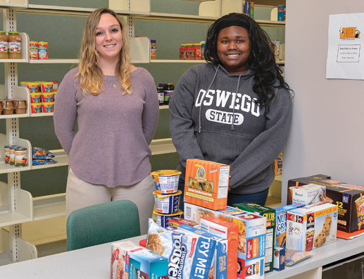 MEETING NEEDS — Shown with some of the nonperishable foods available to SUNY Oswego students in need at the new Students Helping Oz Peers (S.H.O.P.) pantry are volunteers Amanda Sehres ’16 M’17 (left), a school counseling major and a graduate assistant student coordinator of the project, and Rachel McGriff ’14 M’17, a graduate assistant at the Counseling Services Center and a mental health counseling major. The pantry opened in Room 3 in Penfield Library’s basement in October and offers food, toiletries and winter clothing to students in need.